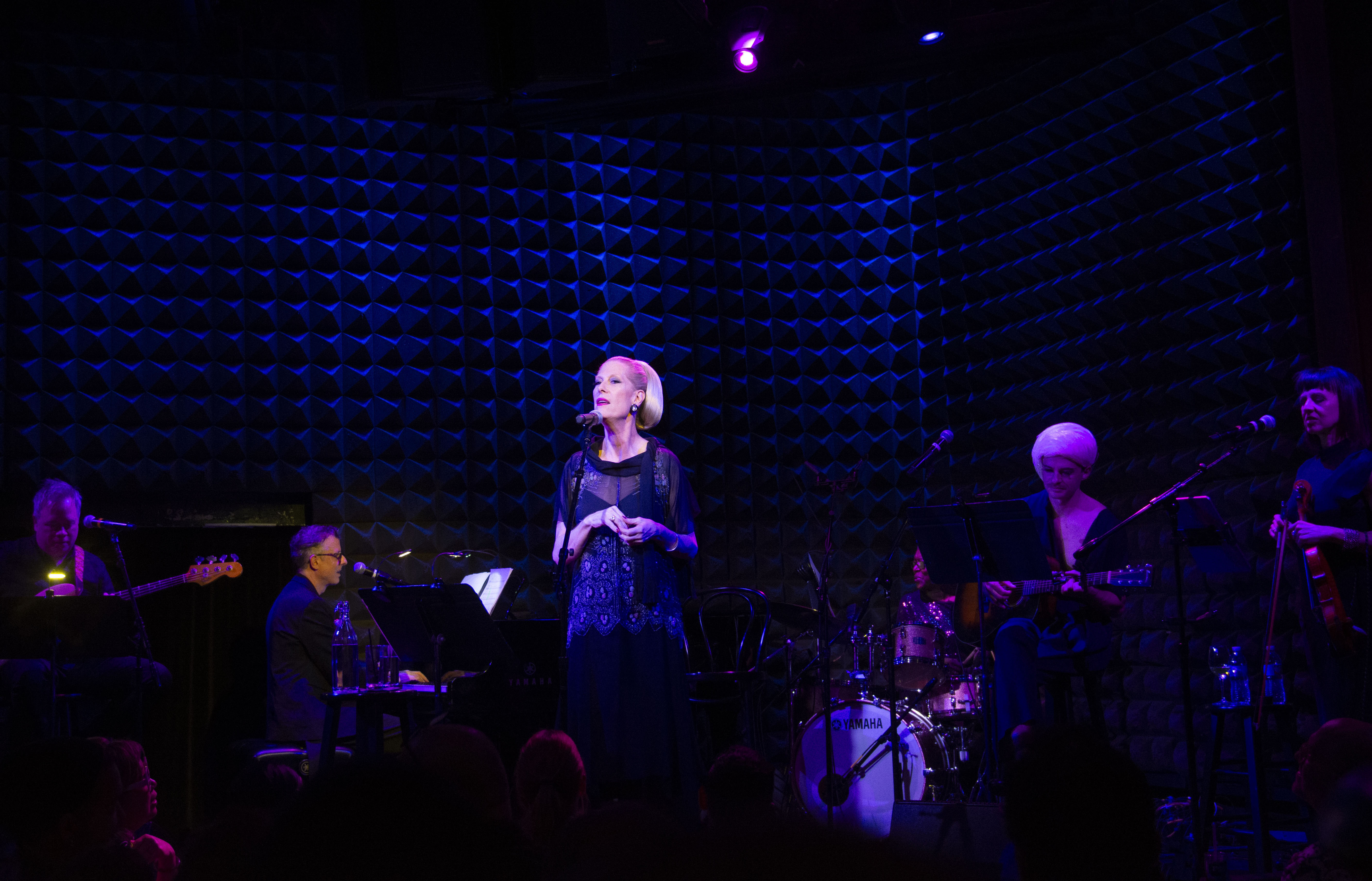 A wide shot of Justin Vivian Bond singing on stage with their band behind them.