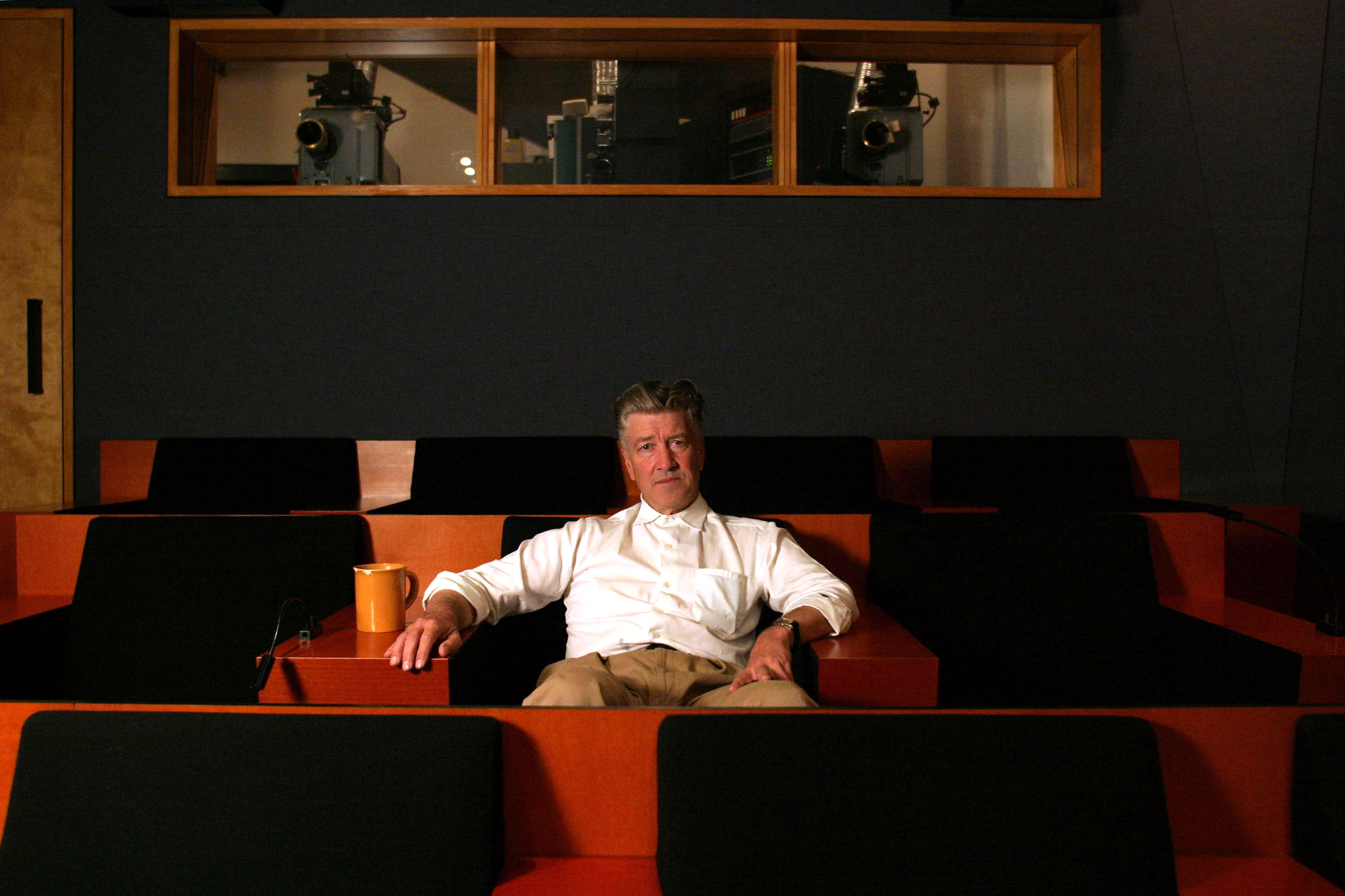 David Lynch sits in a screening room alone on a red seat looking at the camera.