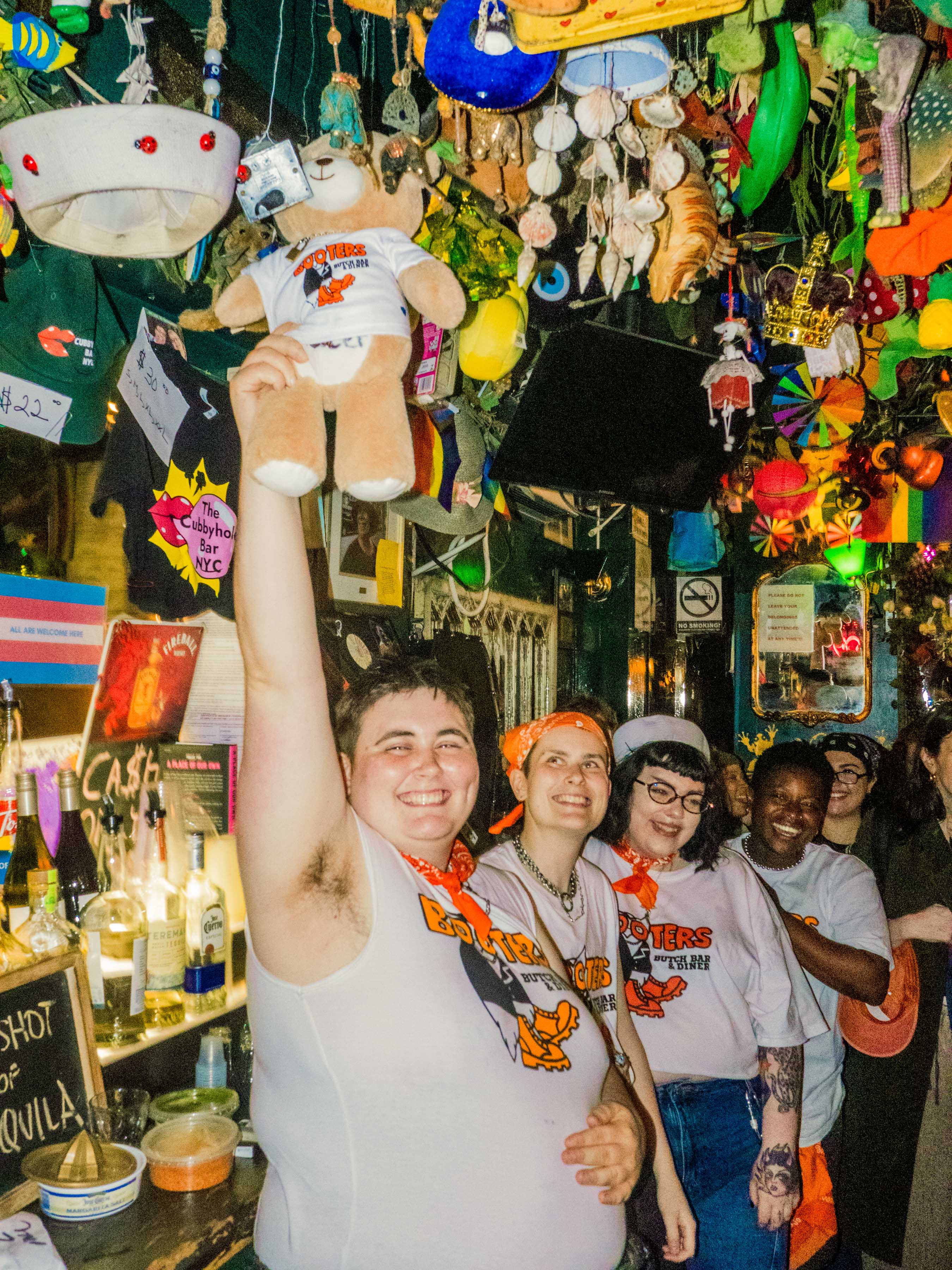 several people, including members of the Booters organizing team, all wearing Booters apparel. The person in the front is holding up a teddy bear with a Booters shirt. They're in a gay bar in NYC with objects hanging from the ceiling.