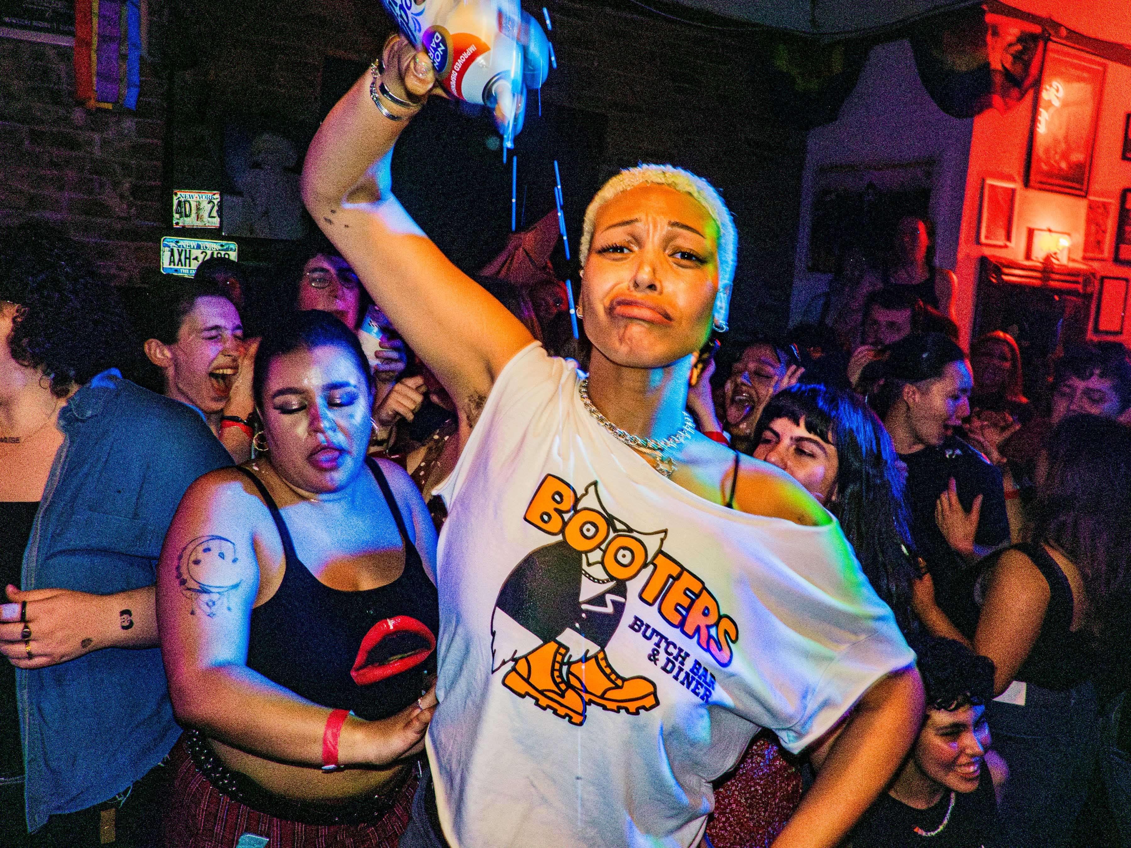 A crowd in the back room at a lesbian bar, with a person wearing a Booters white tee holding a can of whipped cream upside down.