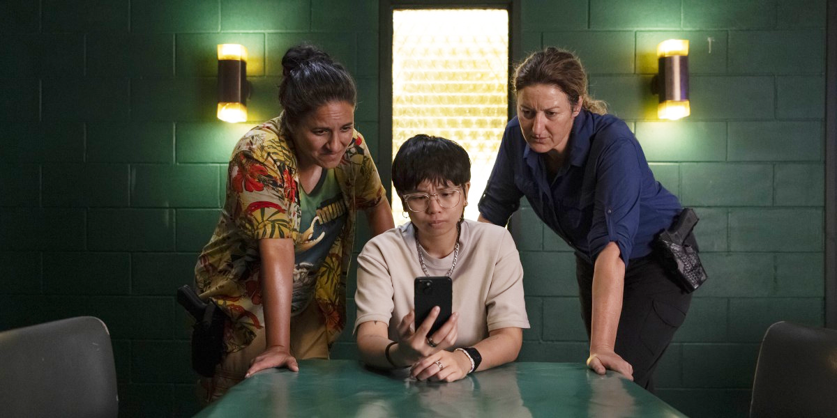 Press shot from Deadloch Season 2 of Eddie and Dulce leaning over Leo, who is looking at something on their phone (courtesy of Amazon MGM Studios)