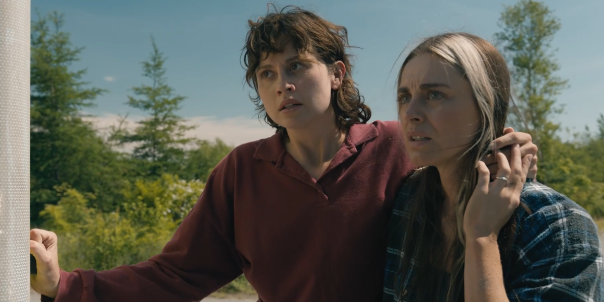 Screenshot from the TV show FROM: Kristi stands with her arm around Mari's neck, Mari holds Kristi's hand that is over her shoulder. They are both looking off screen sternly.
