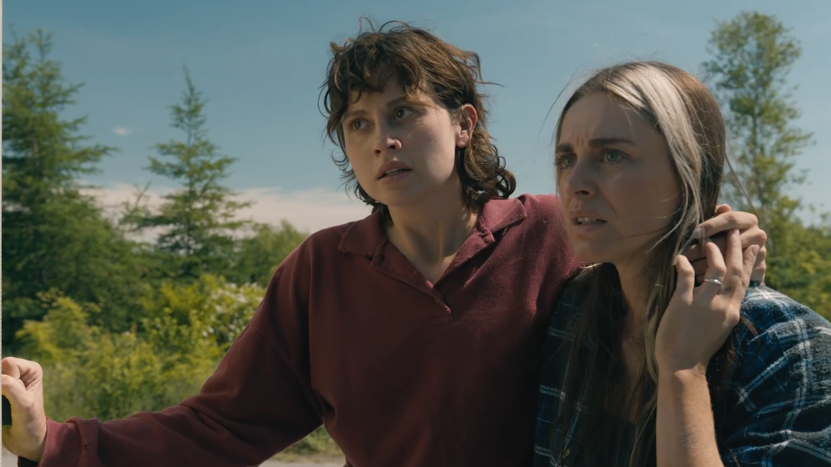 Screenshot from the TV show FROM: Kristi stands with her arm around Mari's neck, Mari holds Kristi's hand that is over her shoulder. They are both looking off screen sternly.