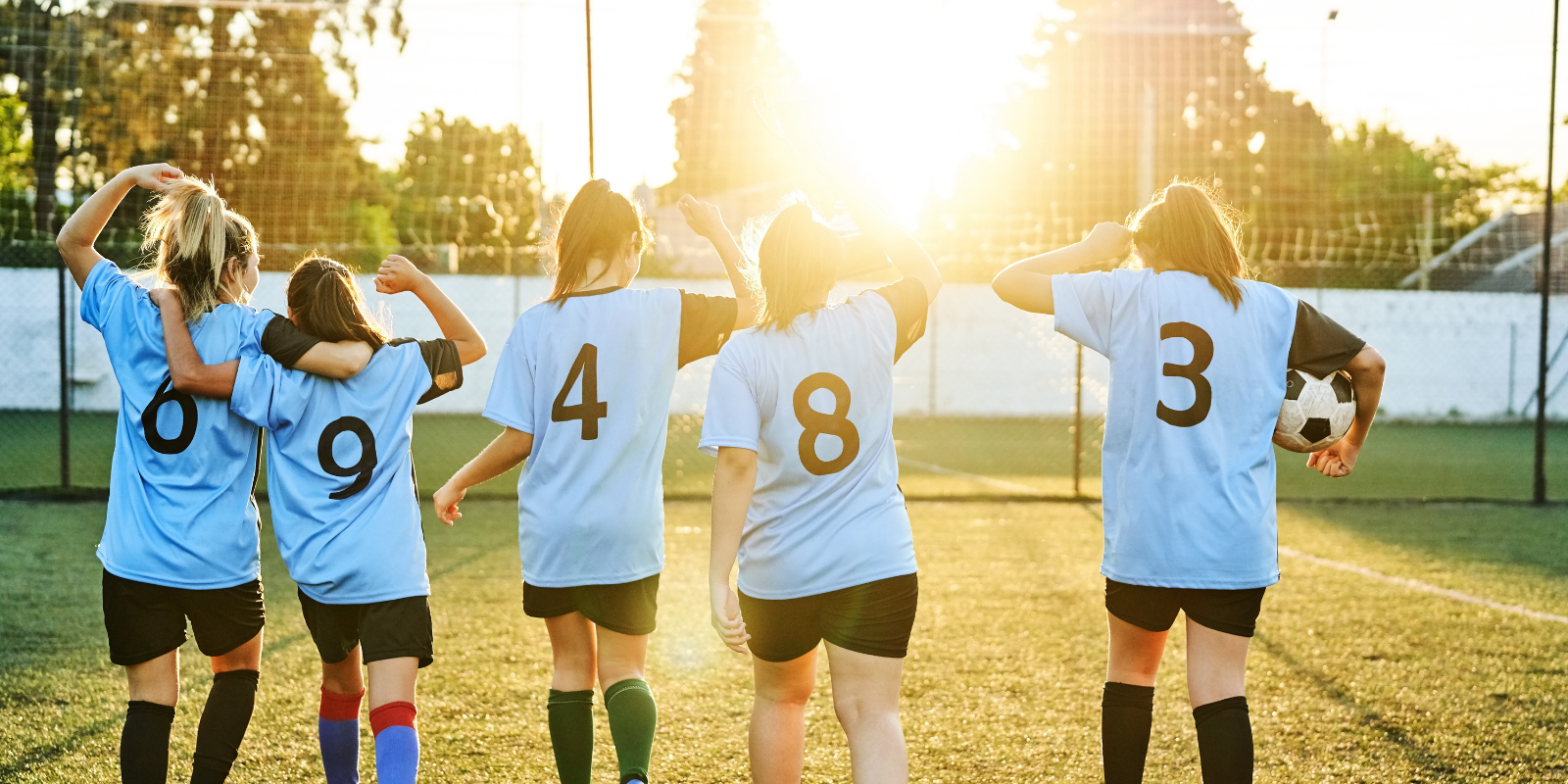 Rear view of girls soccer team walking and celebrating with a trophy on the outdoor field with bright sunlight. Exhilaration and camaraderie as they share their triumphant moment.